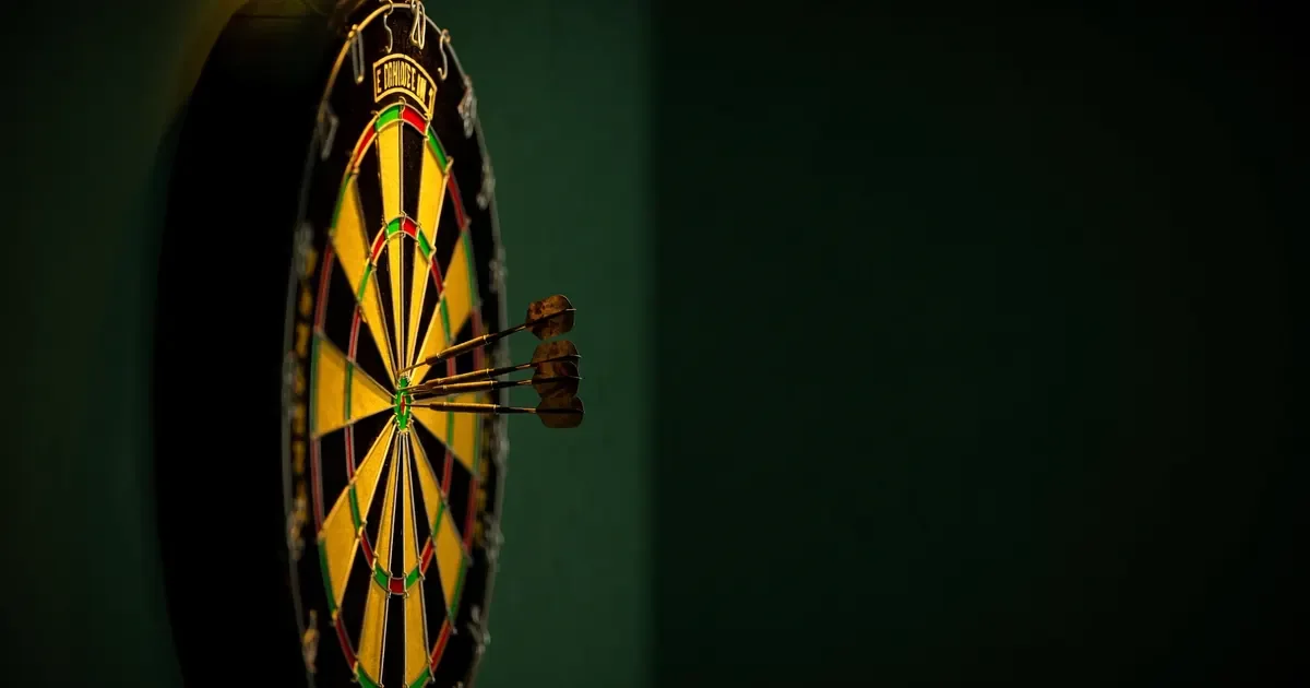 A dartboard on a dark green wall with three darts in the treble twenty